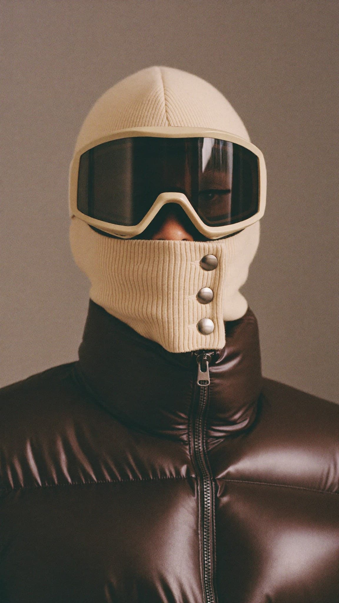 The Knit Balaclava: an ecru rib-knit hood with a goggle in the same ecru and a three-snap gaiter over the nose, worn with a dark brown puffer jacket.