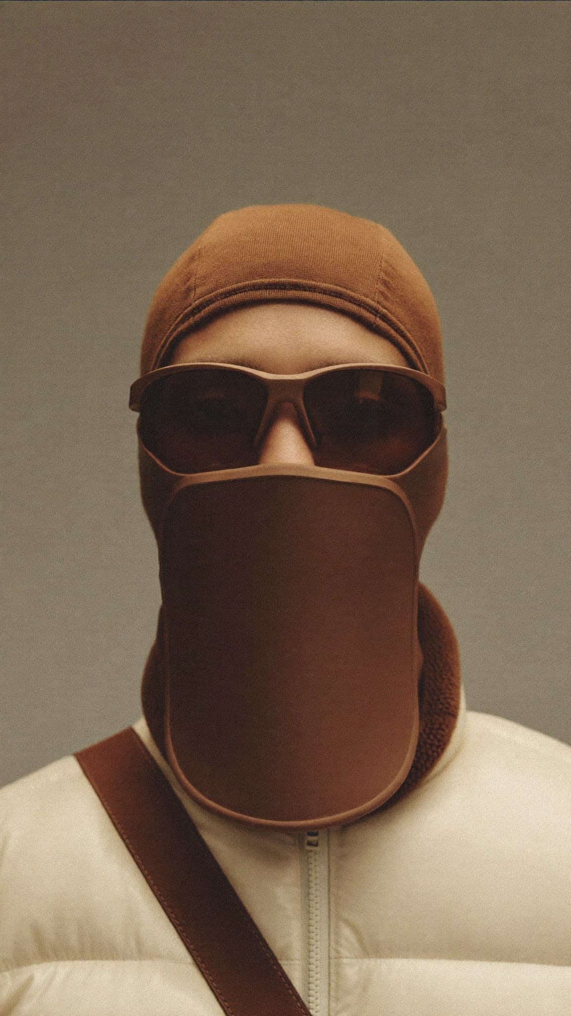 The Shield: a moulded tobacco-brown panel hanging from the frame of a pair of glasses and covering the face to the collar, over a fine-gauge hood, worn with a cream puffer jacket.