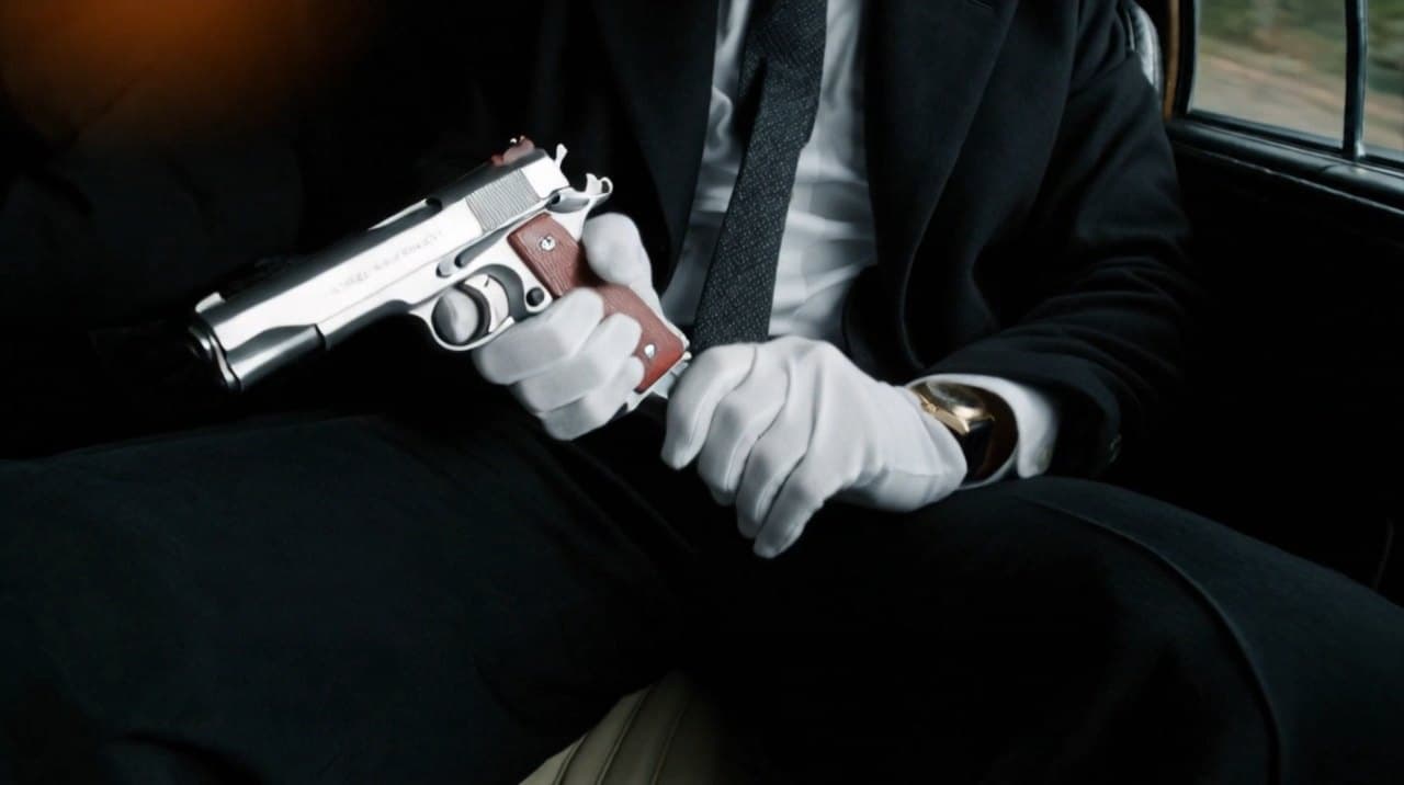 Scene 04: white-gloved hands holding a nickel pistol in the back of the car.